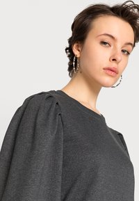 Sweatshirt cinza escuro com mangas bufantes, feito de um tecido macio. O decote redondo e o material texturizado realçam o seu design. Usando brincos de argola prateados.