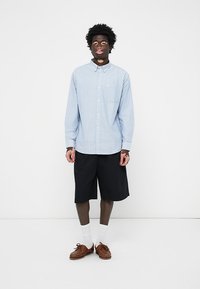 Lichtblauwe gestreepte overhemd met knopen, zwarte shorts, bruine loafers, witte sokken. Het overhemd heeft een enkele borstzak en lange mouwen.