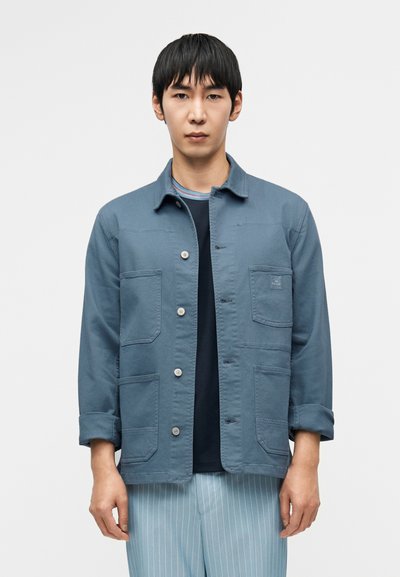 Veste en coton bleue à manches longues, avec poches sur la poitrine et fermeture à boutons, associée à une chemise foncée et à un pantalon clair à rayures.