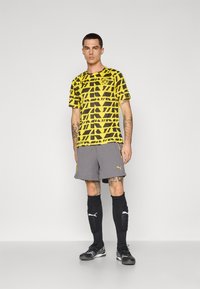 Gul mønstrede fodboldtrøje med sorte geometriske design, parret med grå shorts, sorte sokker og sorte fodboldstøvler.
