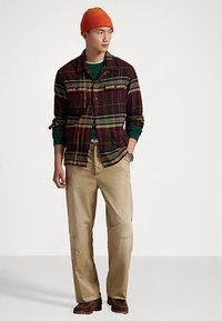 Flanellskjorte i mørk ruter med grønne og brune toner, beige bukser med slitasje, og en oransje strikket lue. Brune sko fullfører antrekket.