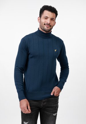 Suéter de cuello alto en color marino con textura acanalada vertical, diseño ajustado y un pequeño logo dorado en el pecho, combinado con jeans de mezclilla oscura.