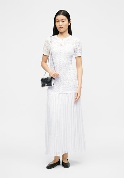Žena v bílé krajkové krátkosrsté halence a plisované maxi sukni, držící černou kabelku přes rameno a obutá v černých balerínkách.