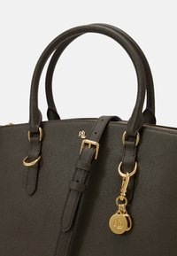 Mörkgrön handväska gjord av texturerat material, med guldfärgad hårdvara, dubbla handtag och en justerbar rem med en dekorativ etikett.