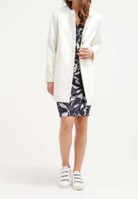 Blazer bianco con un design strutturato, abbinato a un vestito blu navy con stampa di uccelli. Scarpe da ginnastica bianche completano l'outfit.