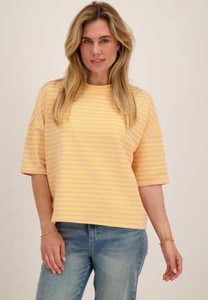 Vrouw met blond haar die een geel en roze gestreept T-shirt met korte mouwen en een blauwe spijkerbroek draagt, staand tegen een effen achtergrond.