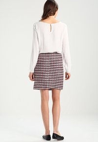 Blusa bianca a maniche lunghe con apertura a chiave sul retro, abbinata a una gonna a fantasia in rosa e nero, e scarpe basse nere con tacco.