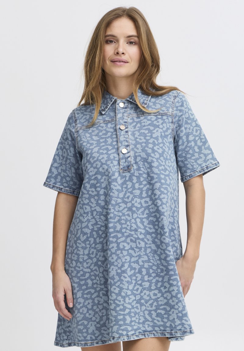 Jeune femme portant une robe en jean bleu à manches courtes et amples, imprimé léopard, avec des boutons à l'avant, debout, une main dans la poche.