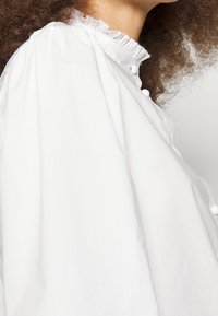 Blusa bianca con colletto alto con bordi arricciati, grandi maniche voluminosi e bottoni rotondi sul davanti. Tessuto dalla superficie liscia.