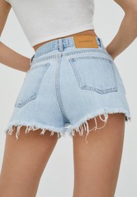 Pantalones cortos de mezclilla azul claro con dobladillo deshilachado, cinco bolsillos y una etiqueta de marca de cuero marrón en la cintura. Corte clásico con un diseño ligeramente ajustado.