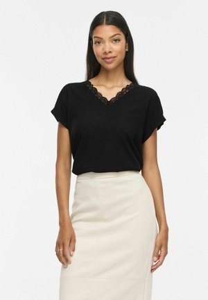 Vrouw met lang donker haar die een zwart topje met V-hals en kantafwerking draagt, en een beige rok met hoge taille, tegen een effen witte achtergrond.