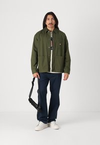 Veste boutonnée vert olive avec deux poches avant, portée sur un t-shirt blanc, associée à un pantalon marine et des baskets blanches. Sac à épaule noir.