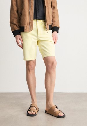 Shorts di denim gialli con taglio dritto, abbinati a sandali slip-on marroni con suola nera e design a doppia cinghia. Giacca bomber marrone sopra.