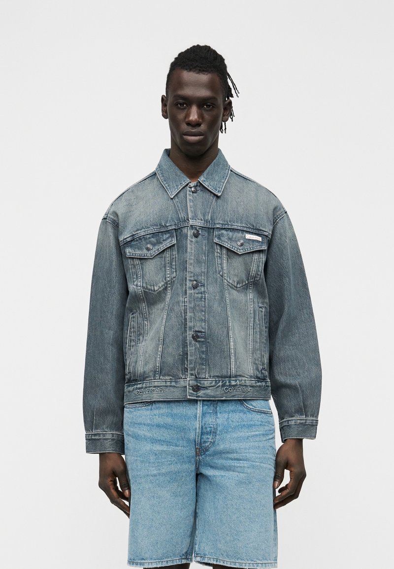 Άνδρας που φοράει ξεθωριασμένο τζιν μπουφάν με τσεπάκια με κουμπιά και ανοιχτόχρωμα τζιν σορτς, στέκεται μπροστά σε απλό λευκό φόντο.