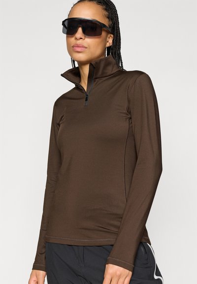 Καφέ μακρυμάνικη μπλούζα με μισό φερμουάρ στον γιακά, που διαθέτει λεία υφή και πλευρικές ραφές. Φοριέται με σκούρες αθλητικές παντελόνες και oversized γυαλιά ηλίου.