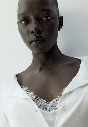 Nahaufnahme einer dunkelhäutigen Frau mit kurzen Haaren, die ein weißes, geknöpftes Oberteil über einem weißen, mit Spitze besetzten Camisole vor einem einfarbigen Hintergrund trägt.