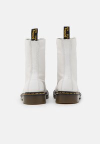 Botas de tornozelo em couro branco com uma textura suave, puxadores com logo amarelo e preto, e costuras contrastantes na sola. Sola plana de borracha durável.