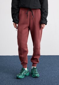 Calças de treino bordeaux com um cós elástico e detalhes de cordão, combinadas com sapatilhas de desporto verdes. O tecido parece suave com uma textura lisa.