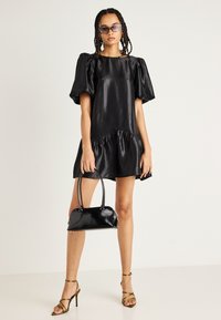 Μαύρο κοντό φόρεμα με φουσκωτά μανίκια και βολάν στο τελείωμα. Το μοντέλο κρατάει μια μικρή μαύρη τσάντα και φοράει χρυσές γόβες με λουράκια και έγχρωμα γυαλιά ηλίου.