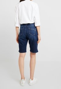 Pantalones cortos de mezclilla en azul oscuro con un corte recto, dobladillo deshilachado y dos bolsillos traseros. Combinados con una camisa blanca de botones y zapatillas blancas.