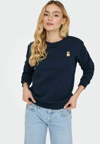 Sweat-shirt bleu marine en tissu doux, avec une petite broderie d'ours en peluche beige sur la poitrine et des poignets et un ourlet côtelés.