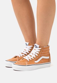 Zapatillas altas de ante en color marrón claro con acentos en blanco, que cuentan con un collar acolchado, cordones blancos y una etiqueta de logo negra en la lengüeta.