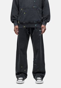 Zwarte oversized hoodie en bijpassende broek met verfspetters in geel, blauw en groen. De broek heeft een elastische tailleband en een relaxte pasvorm.