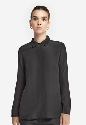 Femme portant une chemise noire à manches longues avec col et boutons, ainsi que des boucles d'oreilles dorées en forme d'anneau, debout devant un fond blanc.