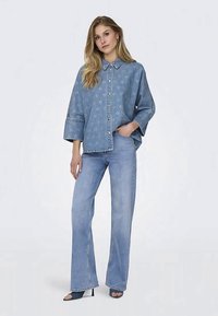 Ženska stoji v svetlobno modrih razširjenih hlačah, modri denim srajci s srčkastim vzorcem in črnimi čevlji z odprtim prstom, roka v žepu, nevtralna ozadja.