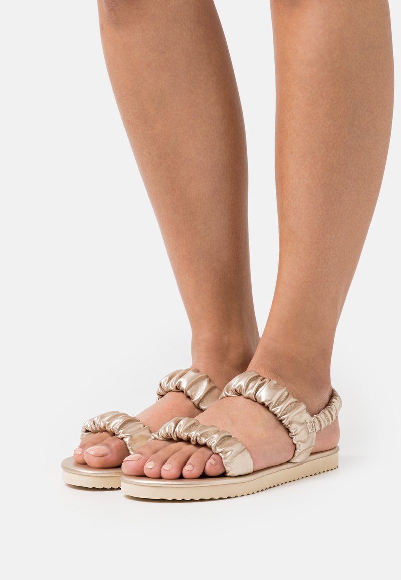 Helle goldene Sandalen mit gerafftem Riemendesign, flacher Sohle und strukturierten Details. Offene Zehenform mit minimalistischem und modernem ästhetischen Ansatz.