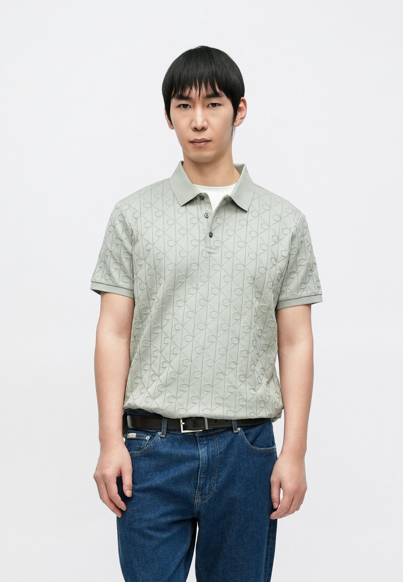 Ανοιχτό πράσινο πόλο μπλουζάκι με υφασμένο διαμάντι σχέδιο, κοντά μανίκια και αντιθετικό γιακά, συνδυασμένο με σκούρο μπλε τζιν και μαύρη ζώνη.