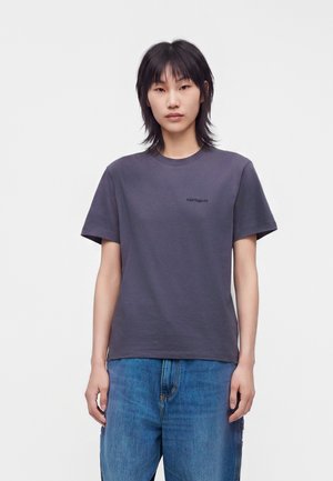 Giovane con capelli neri che indossa una maglietta grigia scura semplice e jeans blu, in piedi davanti a uno sfondo bianco uniforme.