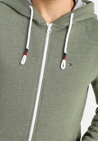 Grüne Zip-Hoodie mit grauem Innenfutter, ausgestattet mit weißen Kordeln mit roten und marineblauen Akzenten sowie einem kleinen Logo auf der Brust. Weicher Stoff.