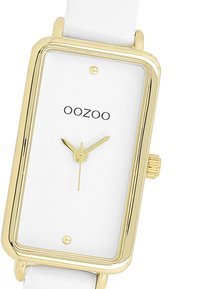 Montre rectangulaire avec un cadran blanc, des accents dorés et des aiguilles dorées fines. Équipée d'un bracelet en cuir blanc et d'un design simple et minimaliste.