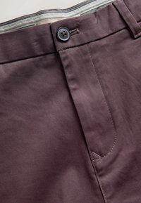 Dunkellila Baumwollhosen mit flachem Vorderteil, ausgestattet mit einem Knopfverschluss und seitlicher Tasche. Glatte Textur mit sichtbaren Nähten.