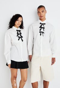 Hvite skjorter med svarte bånddetaljer. En skjorte har flere sløyfer; den andre har en enkelt sløyfe. Inkluderer en svart plissert skjørt og beige shorts.