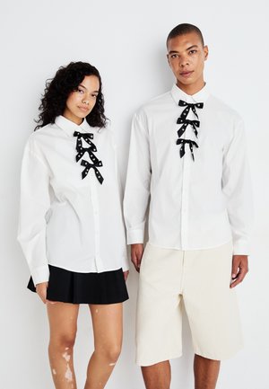 White button-up shirts with black ribbon accents. One shirt features multiple bow ties; the other has a single bow. Includes a black pleated skirt and beige shorts.