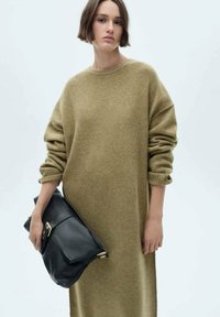 Abito maglione oversize verde oliva con polsini a coste, realizzato in materiale morbido. La figura tiene in mano una borsa in pelle nera con dettagli dorati.