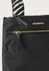 Bolso Gioseppo negro con cremallera y logotipo dorados, que cuenta con una correa a rayas de cebra en blanco y negro y tiradores de cremallera de cuero.