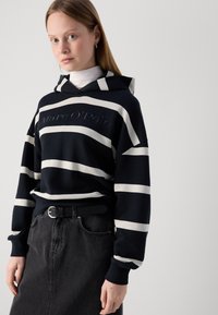 Felpa a righe blu navy e bianco con logo, dotata di tasca a marsupio, polsini a coste e cappuccio regolabile, indossata sopra un dolcevita e una gonna di jeans.