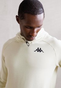 Man wearing off-white hoodie with black logo of two seated figures on chest, looking downward against a plain light background.
