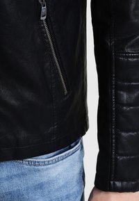 Veste en cuir noir avec une poche zippée, texture matelassée sur la manche et finition lisse. Associée à un jean en denim bleu clair.