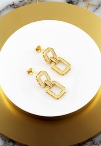 Guldörhängen med länkade rektangulära former, prydda med klara ädelstenar, visade på en cirkulär vit och guldställning.