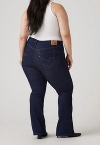 Jeans évasés bleu foncé en denim, dotés de poches arrière avec surpiqûres caractéristiques et d'une étiquette en cuir sur la ceinture. Texture de tissu lisse.