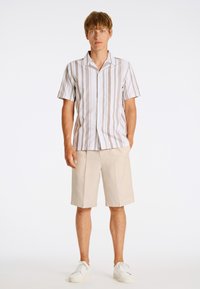 Camicia a maniche corte con bottoni, a strisce verticali beige e bianche, abbinata a pantaloni corti beige e sneakers bianche. Stile semplice e vestibilità rilassata.