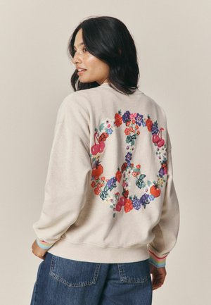 Femeie cu păr închis la culoare, purtând un hanorac bej cu un simbol floral colorat al păcii pe spate și blugi albaștri.