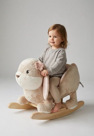 Kleinkind lächelt, sitzt auf einem beigen Plüsch-Kaninchenwippsitz mit Holzschaukel und hält die Ohren des Kaninchens, vor einem neutralen, hellen Hintergrund.
