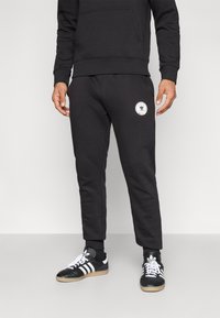 Black sweatpants made of soft material, featuring a fitted design, cuffed hems, and a circular logo on the left thigh. Paired with black and white sneakers.