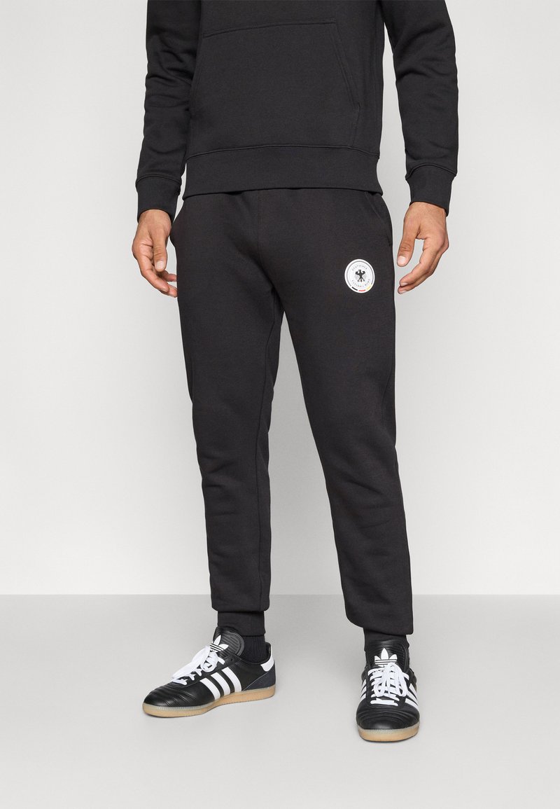 Black sweatpants made of soft material, featuring a fitted design, cuffed hems, and a circular logo on the left thigh. Paired with black and white sneakers.