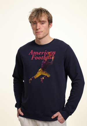 Ung mand iført en navyblå sweatshirt med teksten "American Football" og en illustration af en spiller, der griber en fodbold. Hænderne i lommerne.